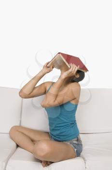Woman sitting on a couch and covering her face with a book