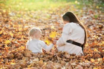 A young mother and little blonde daughter are sitting on fallen leaves in an autumn park, and mom gives her a bouquet of leaves. Mom walks with her daughter in the autumn park.