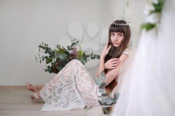 Photo of a young girl in the style of fayn art. A girl in a white translucent dress sits on the floor next to the bed and a bouquet of flowers.