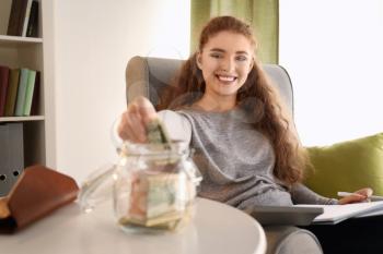 Young woman putting dollar banknote into glass jar indoors. Money savings concept�