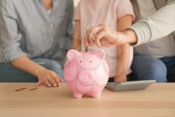 Little girl with her parents  putting coins into piggy bank indoors. Money savings concept�