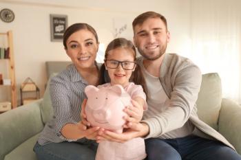 Happy little girl with her parents sitting on sofa and holding piggy bank indoors. Money savings concept�