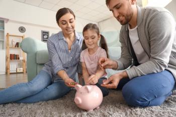 Happy little girl with her parents sitting on carpet and putting coin into piggy bank indoors. Money savings concept�
