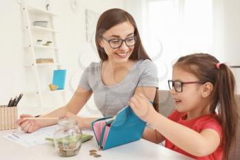 Happy little girl with her mother sitting at table and counting money indoors. Money savings concept�