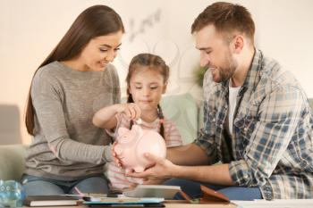 Little girl with her parents sitting on sofa and putting coin into piggy bank indoors. Money savings concept�