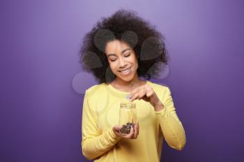 Young African-American woman putting money into glass jar on color background. Savings concept�