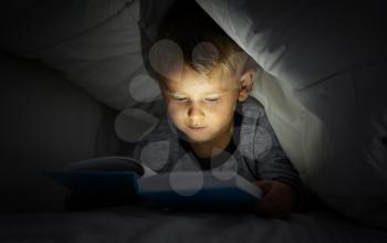 Cute little boy reading book in bed under blanket�