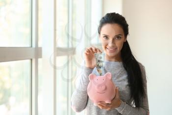Young woman putting money into piggy bank near window�