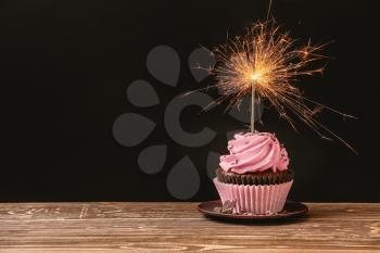 Delicious birthday cupcake with firework candle on table against dark background�