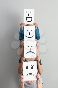 Young women hiding faces behind sheets of paper with drawn emoticons on light background�