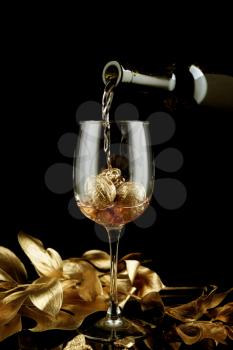 Pouring of pink wine from bottle into glass with gold walnuts on dark background�