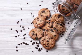 Tasty cookies with chocolate chips and overturned jar on white wooden table�