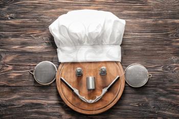 Creative composition with chef's hat, cutting board and kitchen utensils on wooden background�