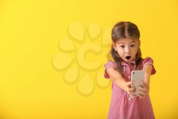 Emotional little girl playing with smartphone on color background�