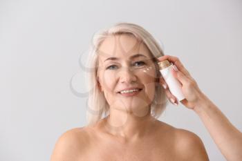 Female hand applying serum onto mature woman's face against light background�
