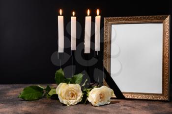 Blank funeral frame, candles and flowers on table against black background�