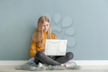 Cute teenage girl with laptop sitting on floor near color wall�