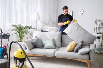 Male janitor cleaning sofa in room�