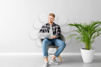 Handsome young man with laptop sitting on chair near light wall�
