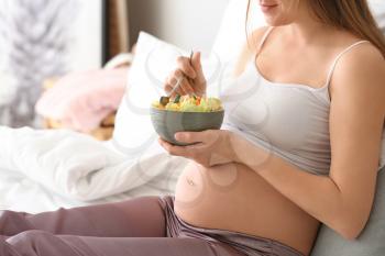 Young pregnant woman eating salad in bedroom�