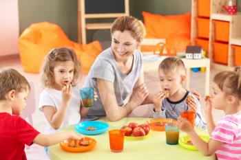 Nursery teacher with cute little children during lunch in kindergarten�