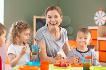 Nursery teacher with cute little children during lunch in kindergarten�