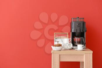 Modern coffee machine, cups and cookies on table against color background�