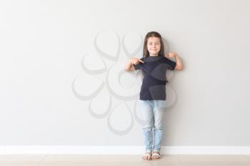 Cute little girl pointing at her t-shirt near light wall�