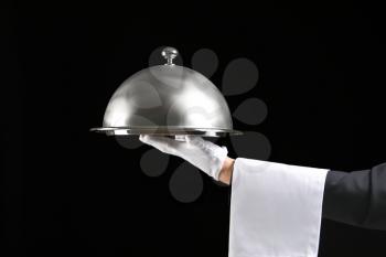 Hand of waiter with tray and cloche on dark background�