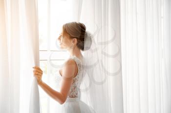 Beautiful young bride near window before wedding ceremony at home�