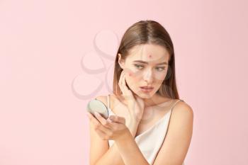 Portrait of young woman with acne problem looking in mirror on color background�