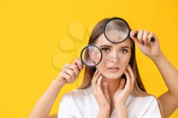 Portrait of young woman with acne problem and hands with magnifying glasses on color background�