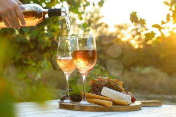 Pouring of tasty wine from bottle into glasses on table with snacks in vineyard�