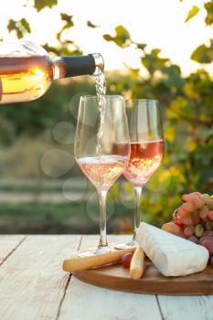 Pouring of tasty wine from bottle into glasses on table with snacks in vineyard�
