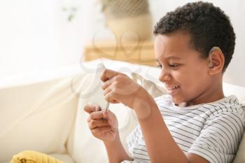Little African-American boy with hearing aid using mobile phone at home�
