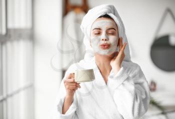 Beautiful young woman with facial mask drinking coffee in bathroom�
