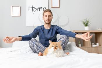 Young man with cute cat meditating in bedroom�