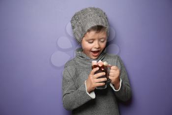 Cute little boy in warm clothes with glass cup of hot chocolate on color background�