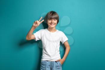 Smiling little boy in t-shirt showing Victory gesture on color background�