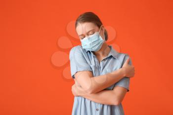 Young woman in medical mask embracing herself on color background�