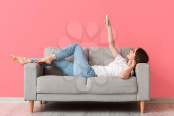 Young woman with air conditioner remote control lying on sofa near color wall�