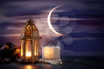 Muslim lamp, glass of milk and tasbih with dates on table against night sky�