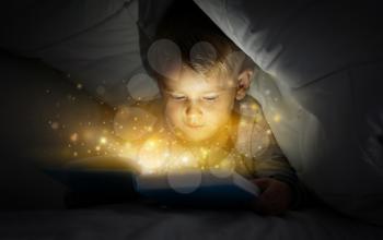 Cute little boy reading magic book in bed under blanket�