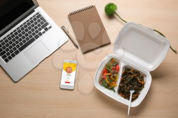 Cell phone with open page of food delivery site on screen, laptop and takeout meal on wooden table�