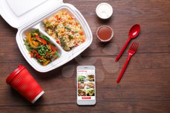 Cell phone with open page of food delivery site on screen and takeout meal on wooden table�