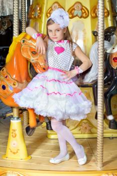 Little beautiful girl in pink and white dress leaning on a carousel pony