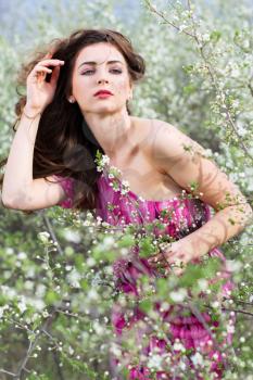 Pretty curly brunette wearing pink dress and posing in blooming garden