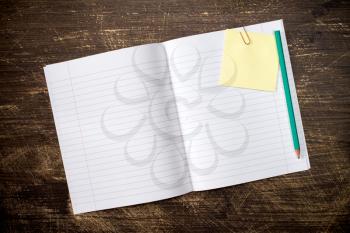 Open exercise-book with sticky note and pencil on wooden background
