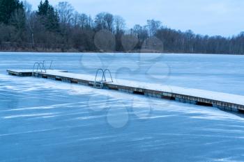 Winter landscape with snow covered pier and frozen lake. Lake covered with thin ice.