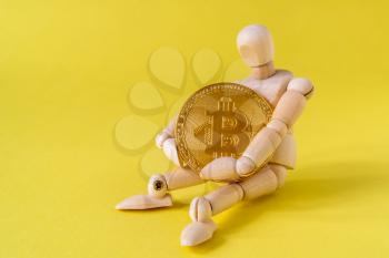 Wooden doll tightly holding Bitcoin while sitting on yellow background